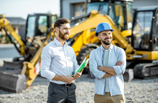 Builder choosing heavy machinery for construction with a sales consultant standing with some documents on the open ground of a shop with special vehicles